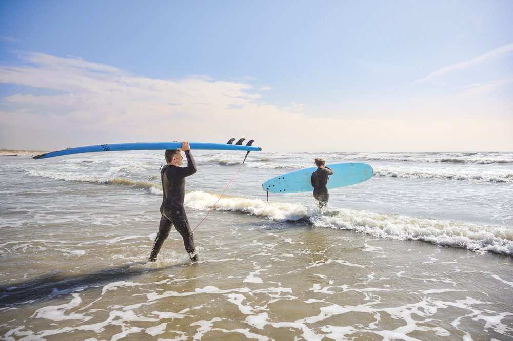A father and daughter, both dressed in black wetsuits, carry blue surfboards into the water at Mustang Island