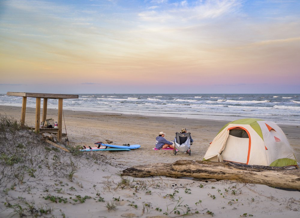 Campers sit next to their tent on the beach, surfboards laying in the sand, staring out at the sun sets over the gulf