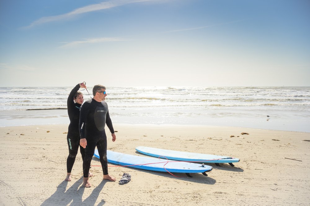 Two surfers in black wetsuits stand on the beach next to their surfboards