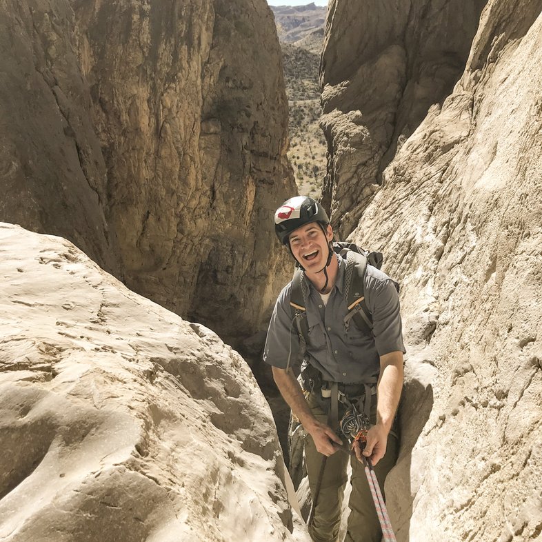 A hiker wearing a helmet and carrying trekking poles stands in the crevice between two large rocks