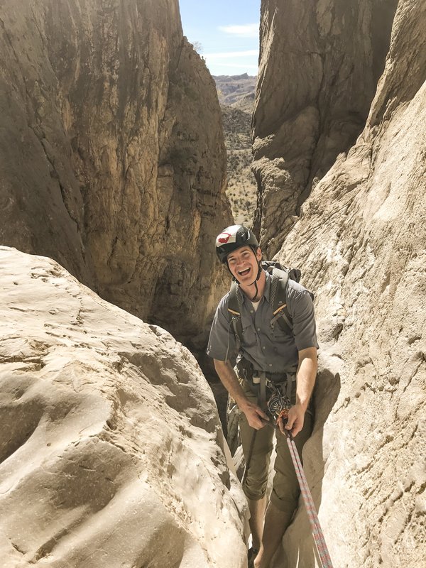 A hiker wearing a helmet and carrying trekking poles stands in the crevice between two large rocks