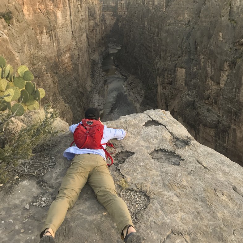 A hiker lays across the surface of the top of a cliff, looking over its edge into the depths below