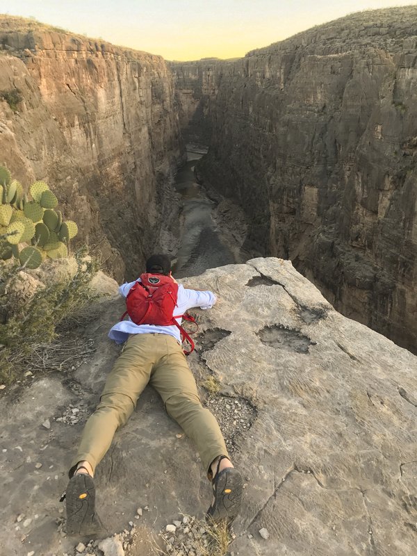 A hiker lays across the surface of the top of a cliff, looking over its edge into the depths below