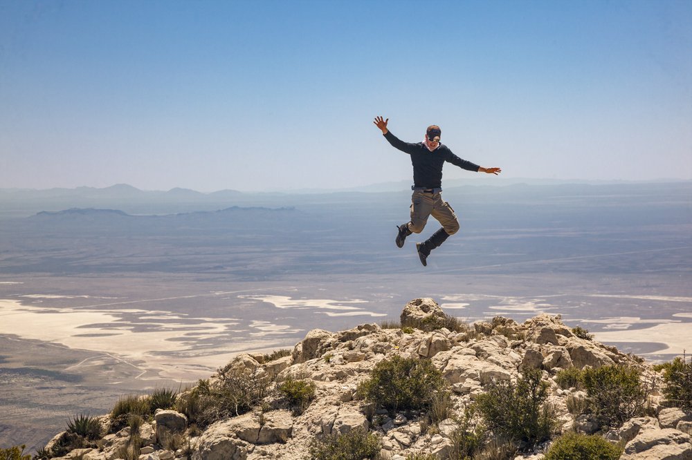 A hiker jumps into the air from the top of a high peak, the valley visible below him in the background