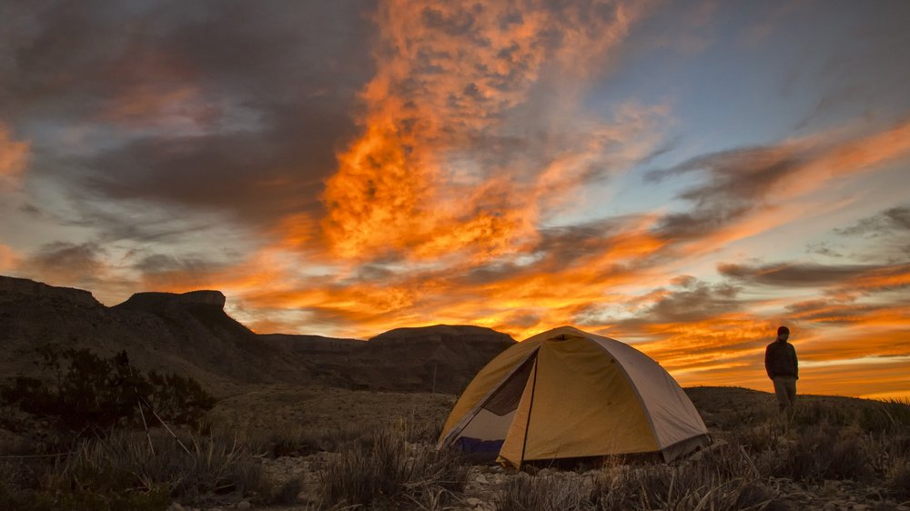 a lone individual stands silhouetted against a golden sunset sky, with a dome tent set up in in front of them.
