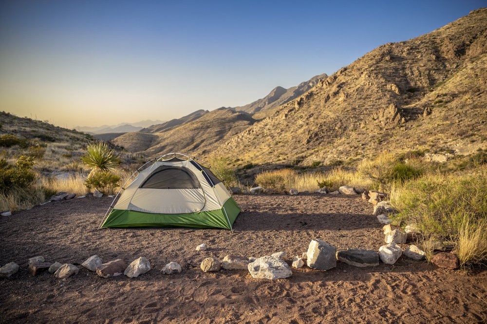 A green and tan dome tent sits surrounded by low rocks at the base of the Franklin Mountains