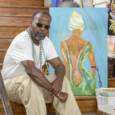 Painter Leon Collins sits on a wooden staircase alongside one of his paintings depicting the back of an African-American woman in a green dress, white headdress and a black chain on her left arm