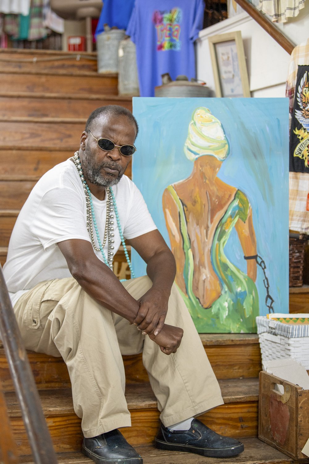 Painter Leon Collins sits on a wooden staircase alongside one of his paintings depicting the back of an African-American woman in a green dress, white headdress and a black chain on her left arm