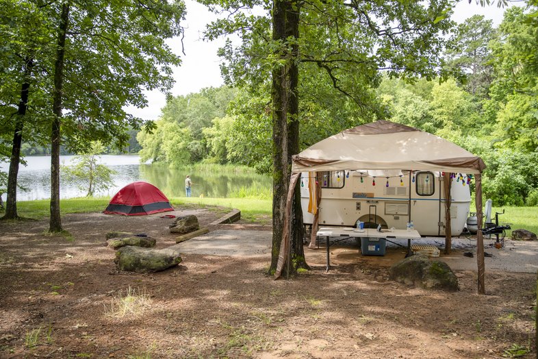 Campsite set up with camper, shade structure and red tent next to lake with man fishing in background