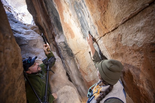 Two archeologists in-between boulders photographing pictographs on rock