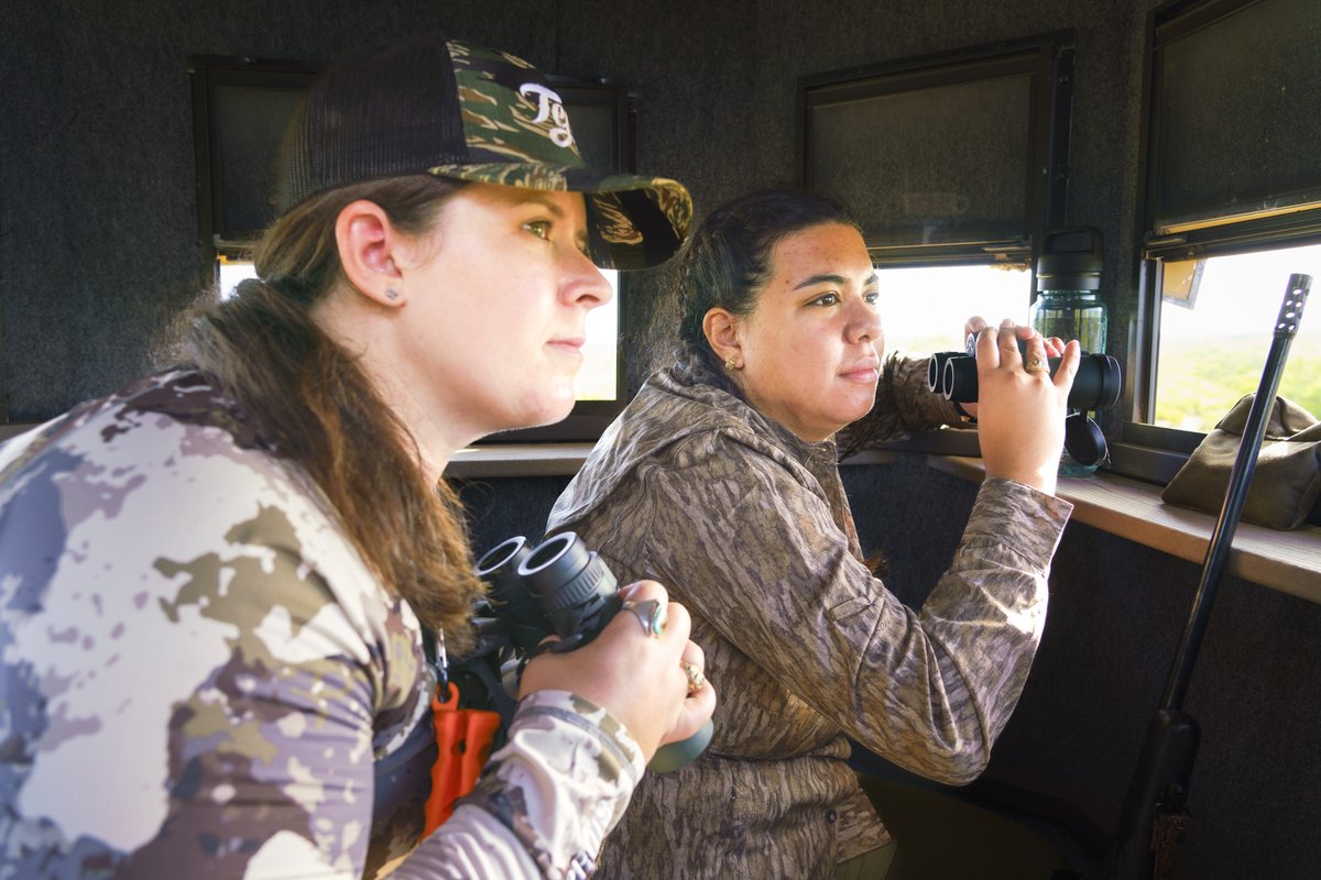 Two female hunters holding binoculars search for deer from inside a deer blind