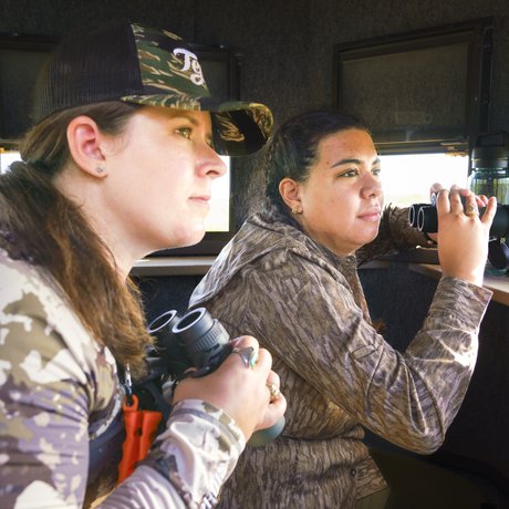 Two female hunters holding binoculars search for deer from inside a deer blind
