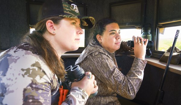 Two female hunters holding binoculars search for deer from inside a deer blind