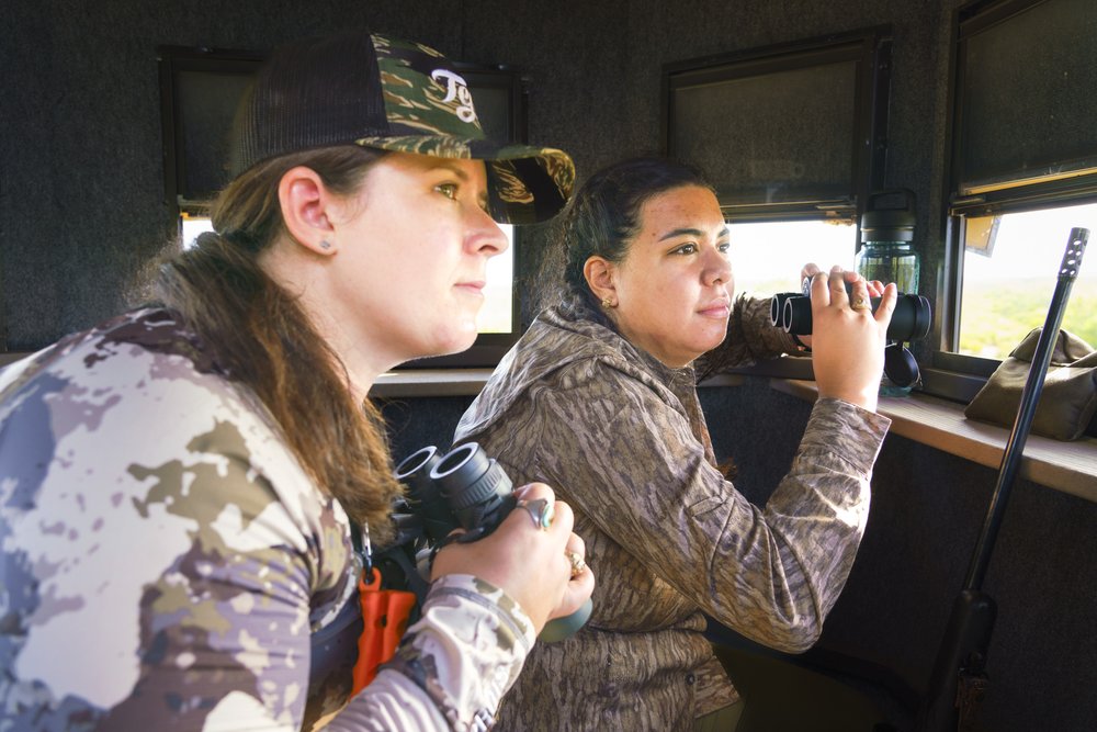 Two female hunters holding binoculars search for deer from inside a deer blind