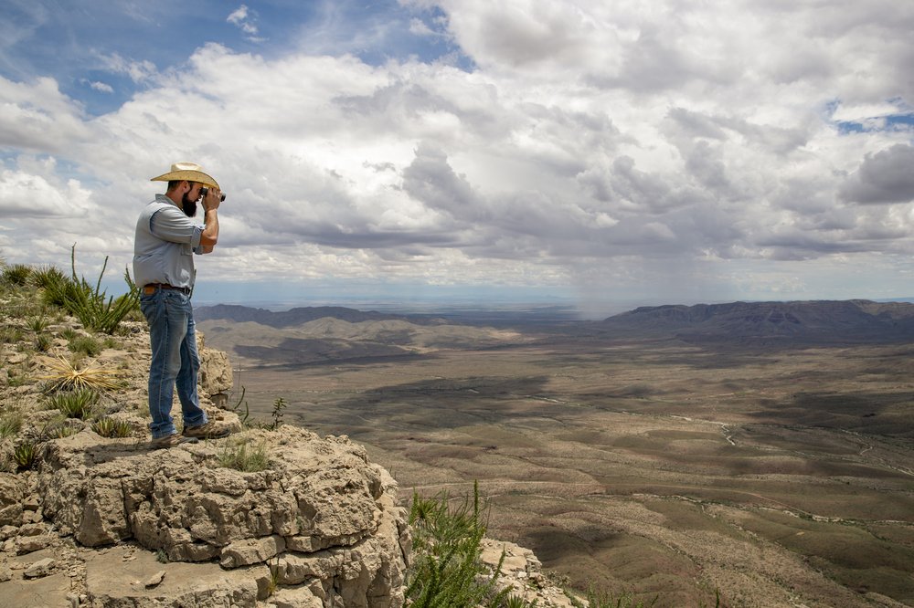 A man standing at the edge of a cliff using binoculars to view the field below.