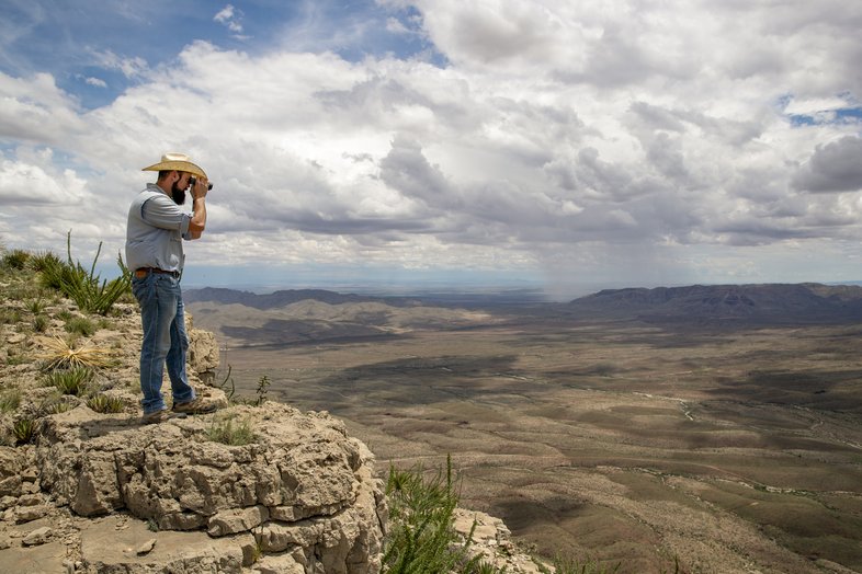 A man standing at the edge of a cliff using binoculars to view the field below.