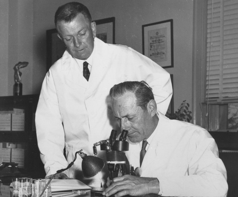 Black and white photo of two men in a laboratory with one looking through a microscope.