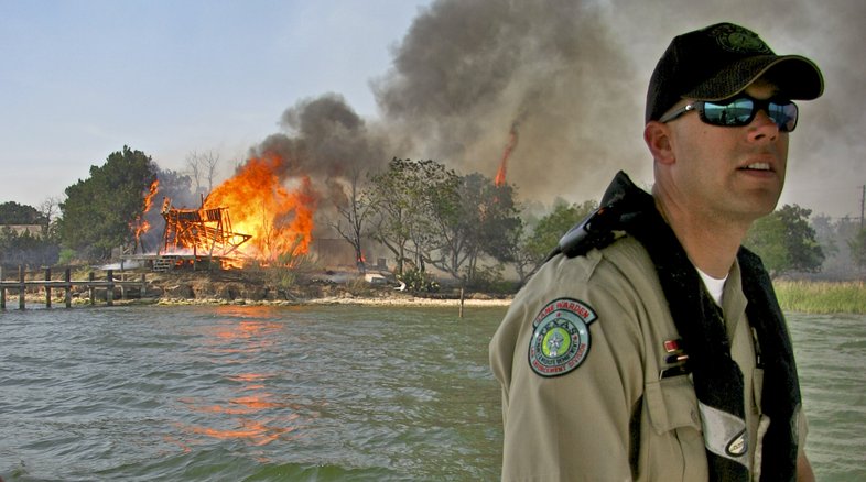 A game warden is on a boat with a fire blazing in the background on shore