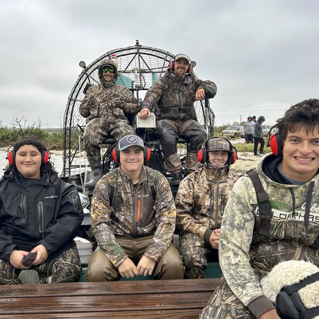 A group of 6 people wearing camo and headphones sitting on a airboat.