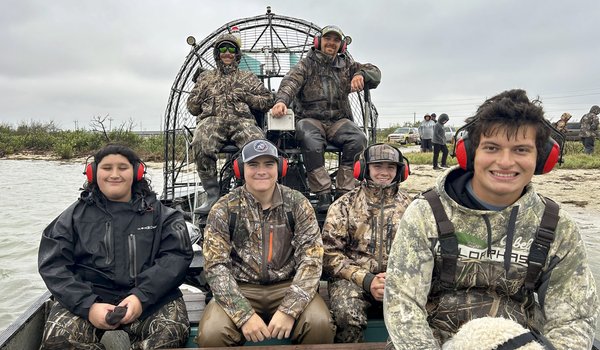 A group of 6 people wearing camo and headphones sitting on a airboat.