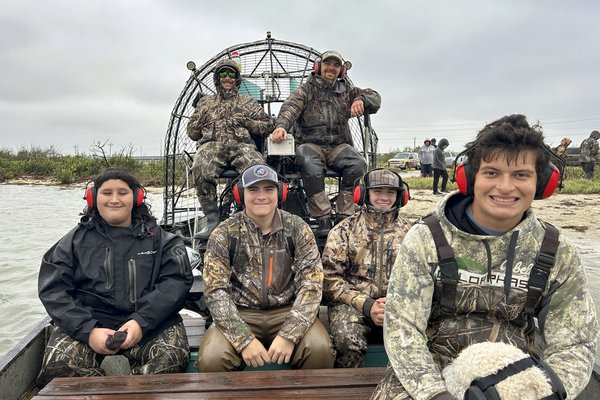 A group of 6 people wearing camo and headphones sitting on a airboat.