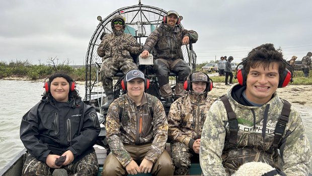 A group of 6 people wearing camo and headphones sitting on a airboat.