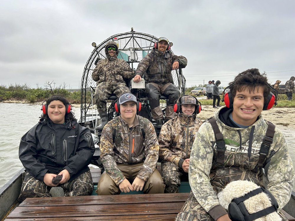 A group of 6 people wearing camo and headphones sitting on a airboat.