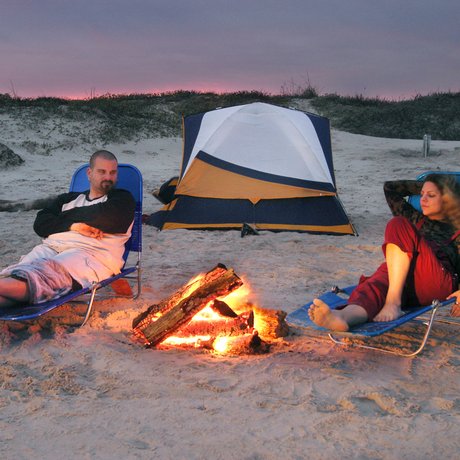 A man and woman relax in lawn chairs on the beach at sunset with a campfire between them and a tent behind them.