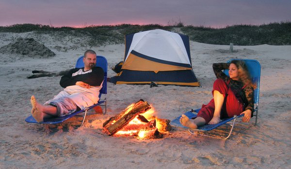A man and woman relax in lawn chairs on the beach at sunset with a campfire between them and a tent behind them.