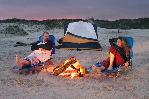 A man and woman relax in lawn chairs on the beach at sunset with a campfire between them and a tent behind them.