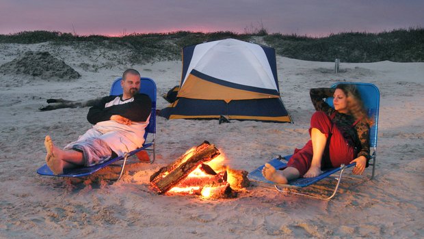 A man and woman relax in lawn chairs on the beach at sunset with a campfire between them and a tent behind them.