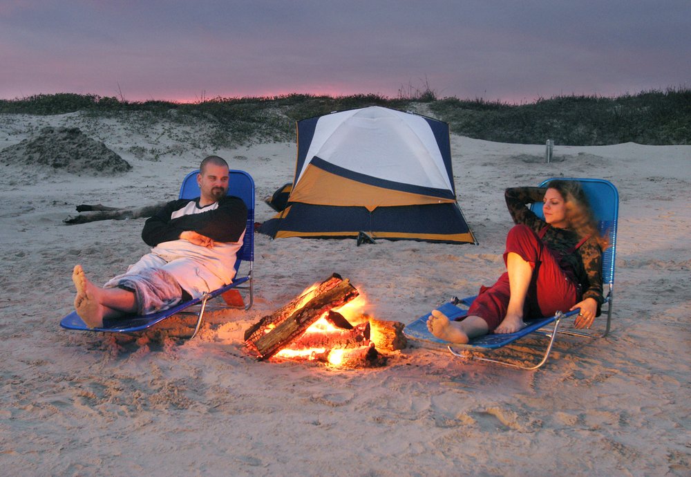 A man and woman relax in lawn chairs on the beach at sunset with a campfire between them and a tent behind them.