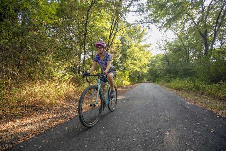 A woman bikes on a paved trail surrounded by trees