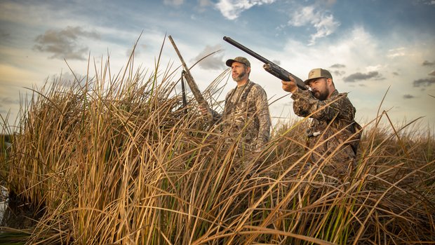 Two men in camoflauge hidden behind tall grass hunting.
