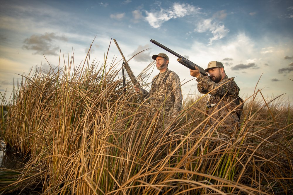 Two men in camoflauge hidden behind tall grass hunting.