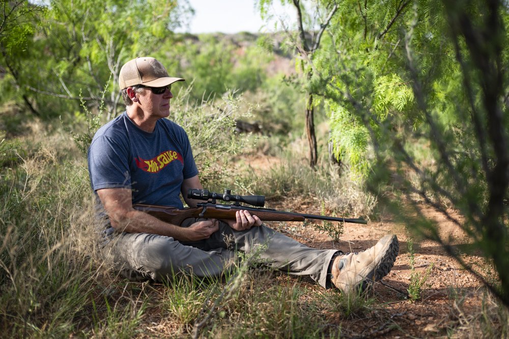 Man sitting on the ground with a gun while hunting invasive species.