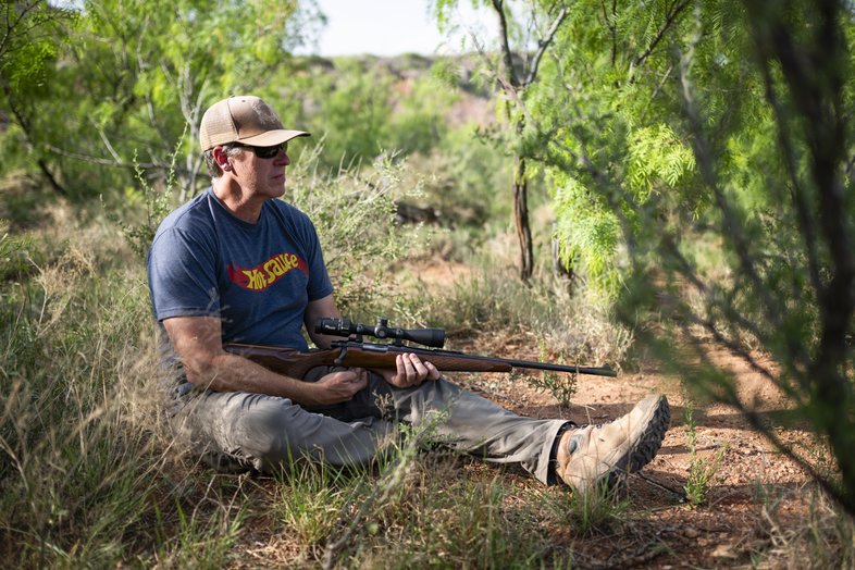 Man sitting on the ground with a gun while hunting invasive species.