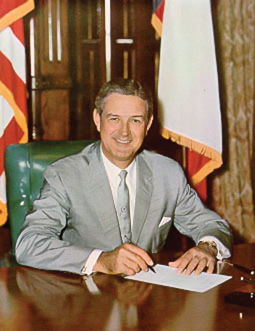 Connally at desk writing with flags behind him