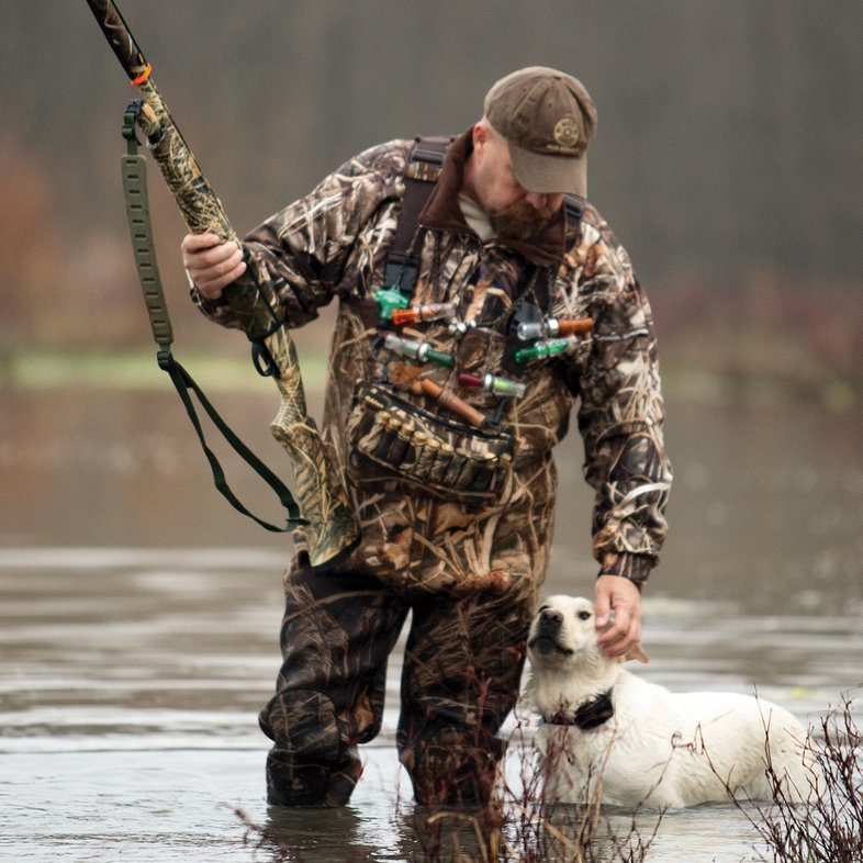 Duck hunter standing water with a gun and a white dog.