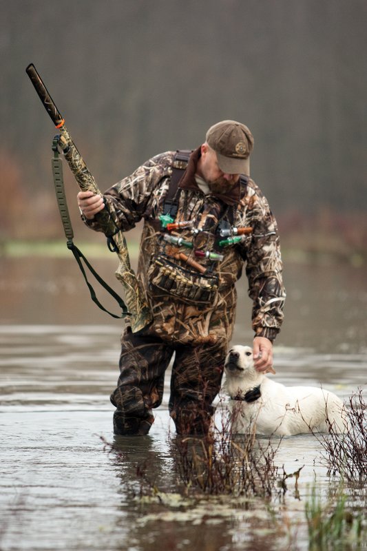 Duck hunter standing water with a gun and a white dog.