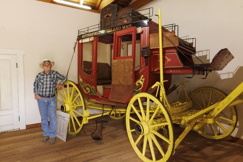 Man standing next to a red stagecoach with yellow wheels at Fort Chadbourne.