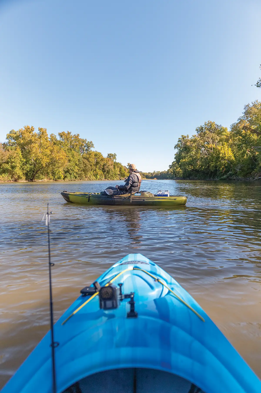 Front of blue kayak being launched into a river at a river access site with a yellow kayak on the river