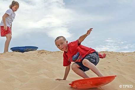 Girl with a blue sled at the top of a sand hill iwth a boy on a red sled surfing down the hill