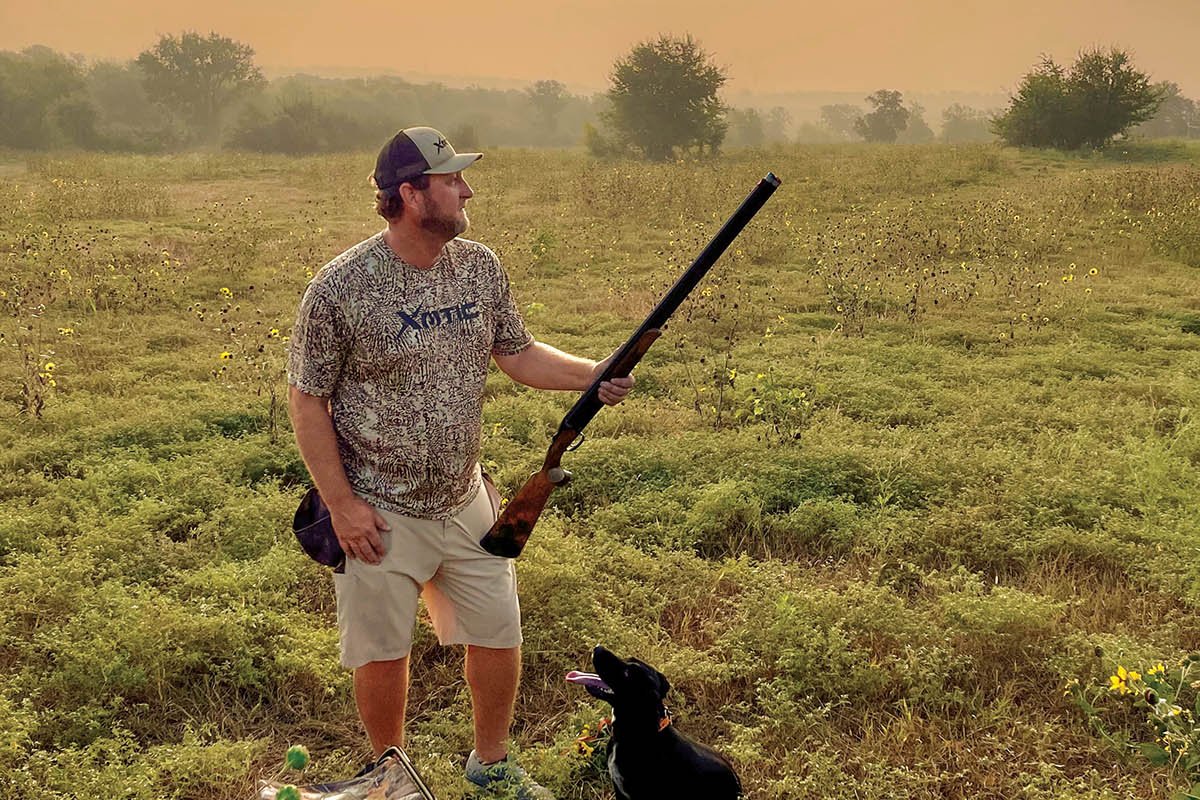 Hunter holding a gun with a black dog at his feet looking up.