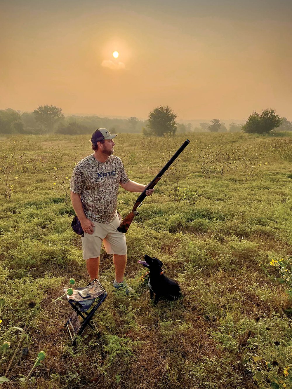 Hunter holding a gun with a black dog at his feet looking up.