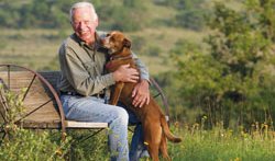 J. David Bamberger siting on a wagon wheel bench in a feild with his dog.