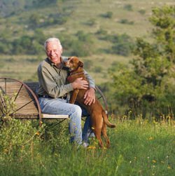 J. David Bamberger siting on a wagon wheel bench in a feild with his dog.