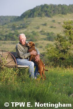 J. David Bamberger siting on a wagon wheel bench in a feild with his dog.