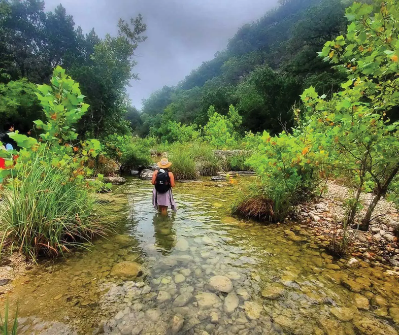 Josei-Gutierrez walking toward a hill in a stream with a rocky bottom and trees surrounding