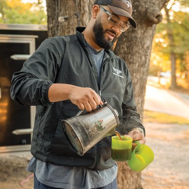 Man in a hat pouring coffee into one mug while holding another at a campsite